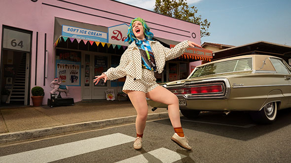 Libby is dancing with arms outstretched on a pedestrian crossing. A vintage car parked on the street in the background outside a store