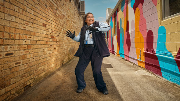 Margie dancing with waving arms in an urban alleyway. A colourful painted wall to one side