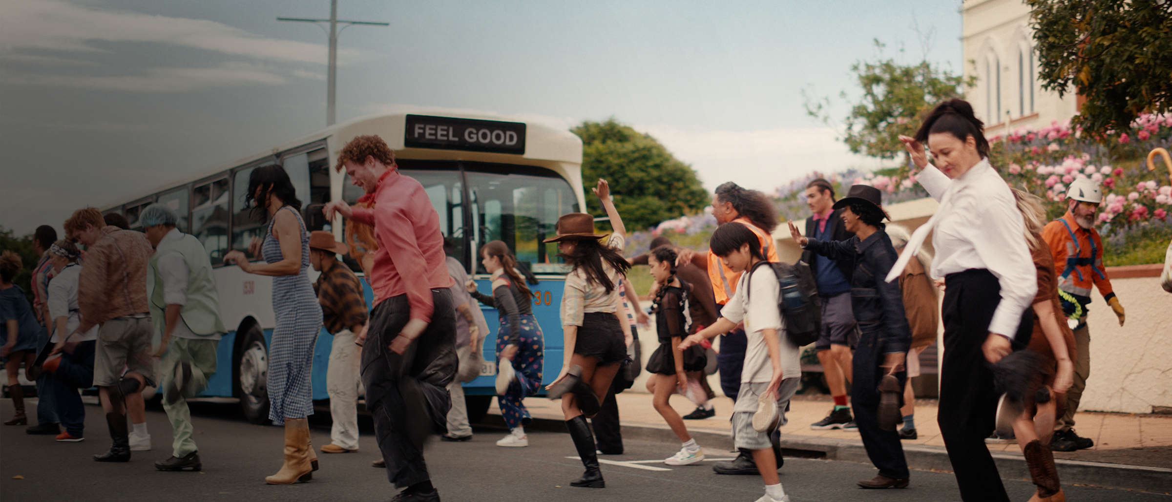 A large group of happy people dancing in the street. A bus with the words "Feel Good" in the background.